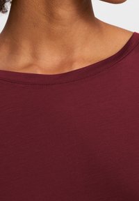 Bordó top s krátkým rukávem, s hladkým povrchem a širokým výstřihem. Obsahuje jemné prošívání kolem límce pro dodatečný detail.