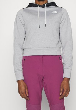 Persoon draagt een lichtgrijze North Face-hoodie met zwarte capuchon en magentakleurige broek, staand tegen een effen achtergrond.