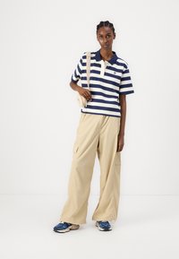 Polo a righe blu e bianche con maniche corte, abbinato a pantaloni cargo beige larghi e sneakers blu, mentre tiene una piccola borsa beige.