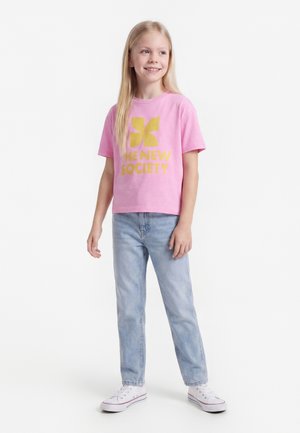 Meisje met lang blond haar, een roze T-shirt met gele tekst en lichtblauwe spijkerbroek dragend, glimlachend en staand tegen een witte achtergrond.