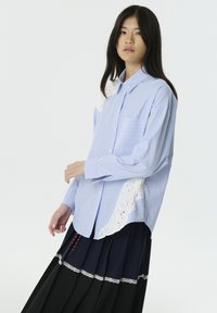 Blauw-witte gestreepte button-up shirt met kantdetails aan de zijkant, voorzien van een borstzak en een ontspannen pasvorm. Gecombineerd met een gelaagde zwarte rok.