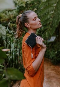 Black textured wallet with a beige zipper and charm, held in a hand. Worn with an orange ribbed garment, set against lush green foliage.