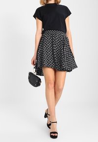 Mujer con blusa negra de manga corta, falda acampanada con estampado en blanco y negro, sandalias de tacón negras y un pequeño bolso negro en la mano.