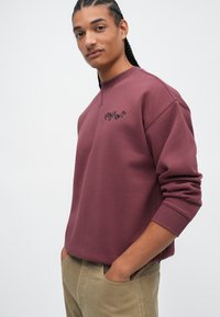 Sweatshirt en coton bordeaux avec col rond et poignets côtelés, présentant un petit logo brodé sur la poitrine. Porté avec un pantalon en velours côtelé beige.