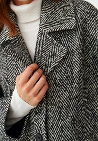 Hånd, der justerer kraven på en sort-hvid høringsbenmønstret frakke, som bæres over en hvid ribbet rullekravesweater.