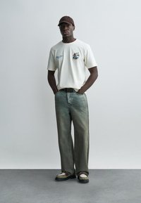 Mees, kes kannab valget graafilise mustriga t-särki, pleekinud laia lõikega pükse, pruuni mütsi ja beeže jalatseid, seisab käed taskutes ühtlase tausta ees.