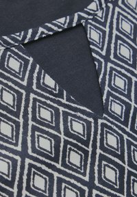 Mörkblått tyg med ett vitt geometriskt diamantmönster. Har en nyckelhålsringning och texturerade detaljer. Standardstickning synlig.