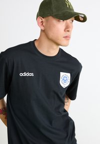 Fekete pamut póló kerek nyakkal, bal oldalon fehér Adidas logóval és kék részletekkel ellátott pajzs logóval a mellkason.