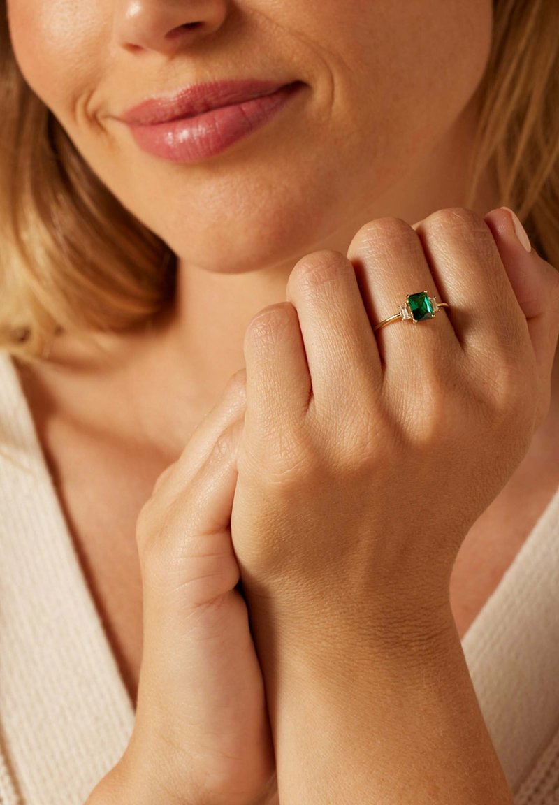 Frau hält ihre Hand nahe ihrem Kinn, trägt einen goldenen Ring mit einem quadratischen grünen Edelstein an ihrem Finger.