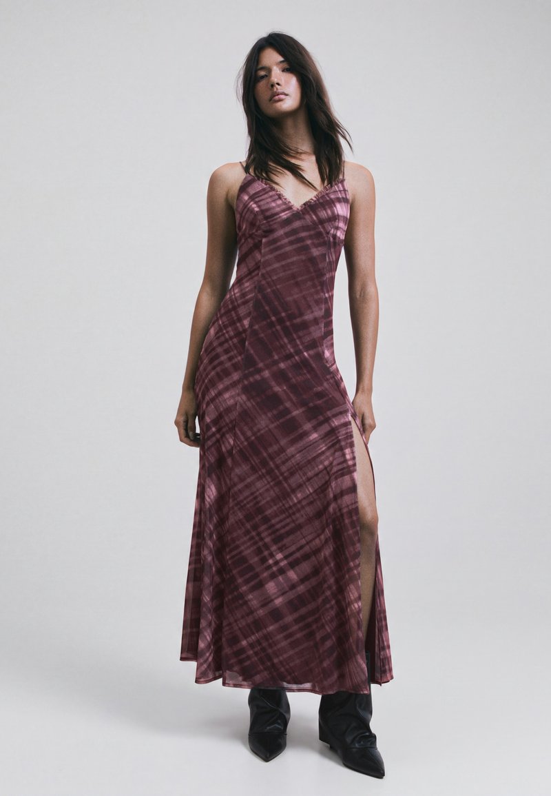 Femme debout portant une robe longue sans manches violette à motifs avec une fente latérale et des bottes noires à bouts pointus sur un fond clair uni.