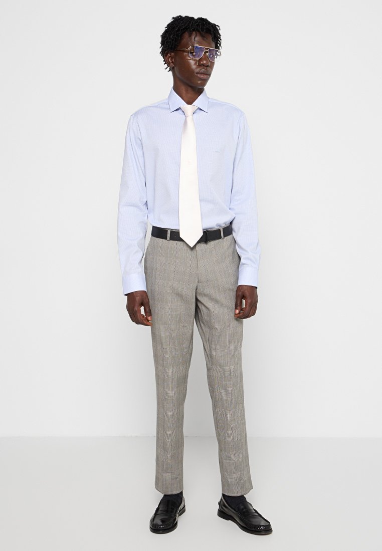 Camisa de vestir a rayas azul claro con una corbata blanca, pantalones de cuadros grises, cinturón negro y zapatos negros brillantes, usados con gafas rectangulares.