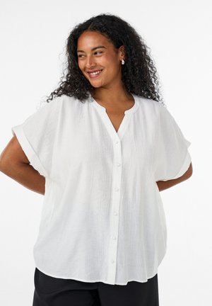 Vrouw met krullend haar die glimlacht, draagt een witte blouse met korte mouwen en knopen en een zwarte broek, handen op de heupen, tegen een witte achtergrond.