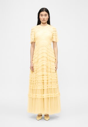 Pitkämustatukkainen nainen, jolla on lattiapitkä vaaleankeltainen kerroksilla oleva tüllimekko, jossa on lyhyet hihat ja teräväkärkiset kengät, seisoo valkoista taustaa vasten.