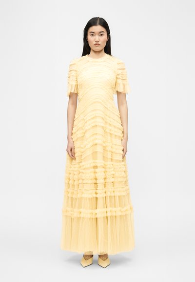 Femme aux longs cheveux noirs portant une robe longue en tulle jaune pâle à volants avec manches courtes et chaussures pointues, posant devant un fond blanc.