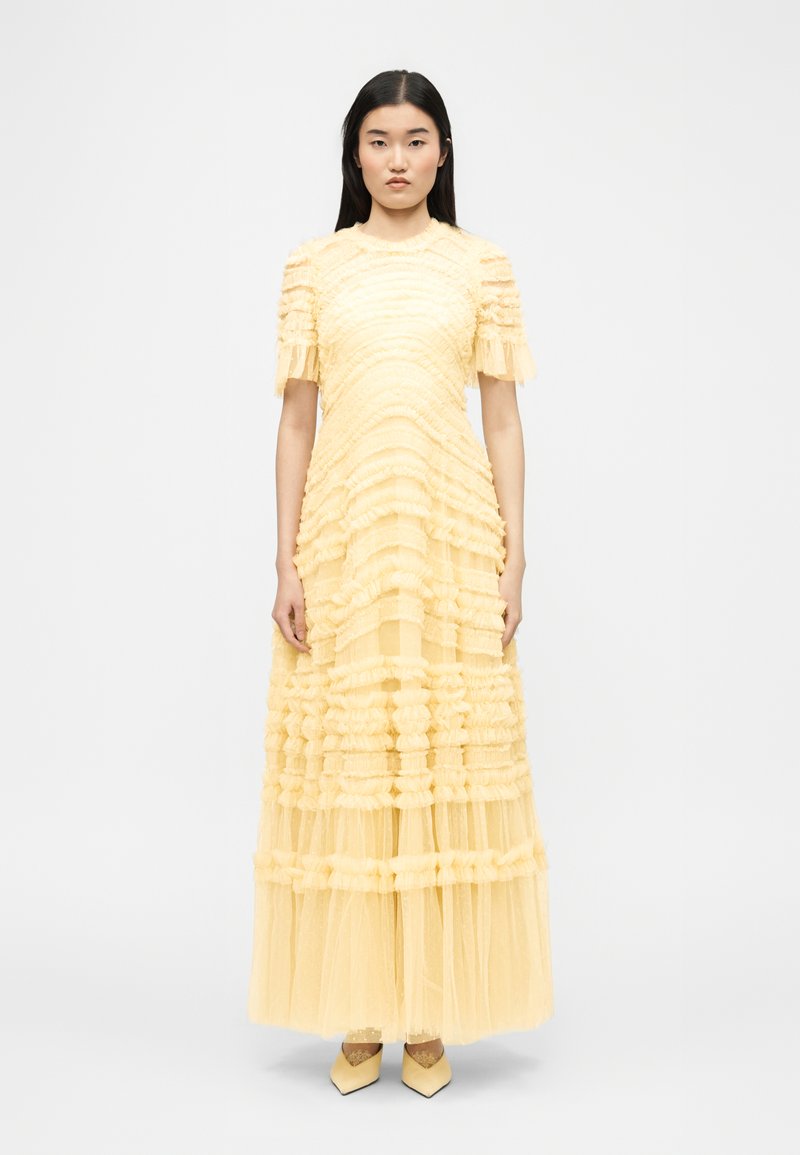 Γυναίκα με μακριά μαύρα μαλλιά φοράει ένα παστέλ κίτρινο φόρεμα από τούλι με βολάν μέχρι το πάτωμα, με κοντά μανίκια και μυτερά παπούτσια, στέκεται μπροστά σε λευκό φόντο.