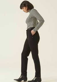 Pantalons noirs fuselés avec un bouton à l'avant, associés à un haut à rayures noir et blanc à manches longues. Le modèle se tient de côté, portant des bottes pointues.