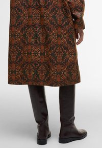Gemusterter Mantel in Dunkelgrün mit orangefarbenem Blumendesign, mit gerader Schnittführung, Knöpfen und kombiniert mit kniehohen braunen Lederstiefeln.