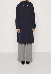 Manteau imperméable bleu marine avec capuche ajustable, taille à cordon de serrage et manches longues. Fermeture éclair à l'avant et poches latérales. Associé à un pantalon gris.