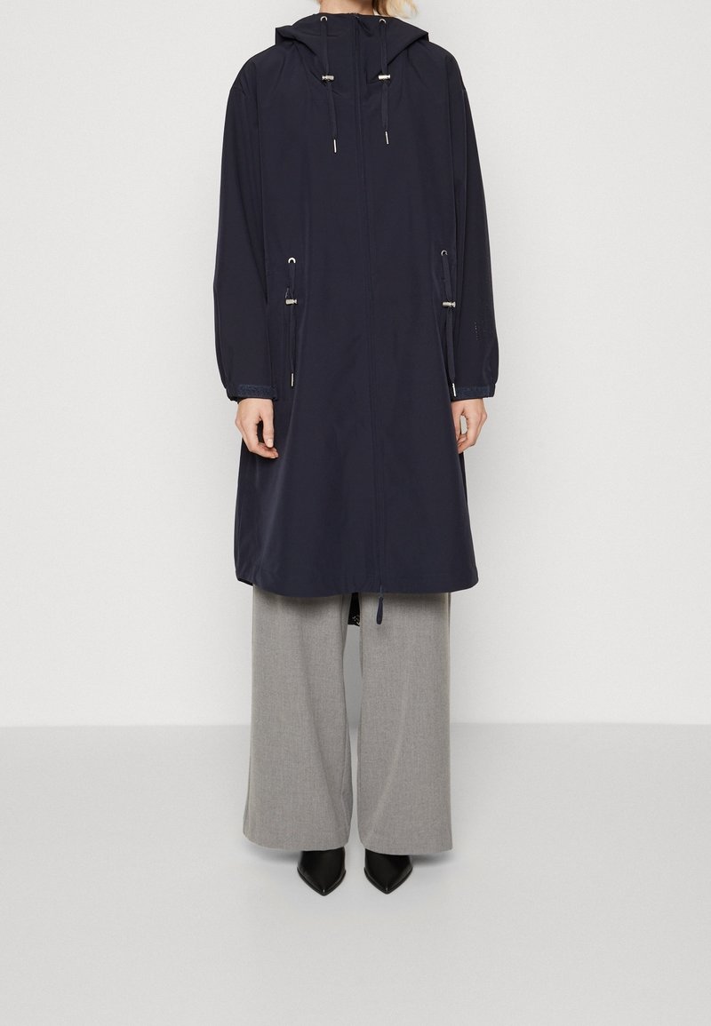 Manteau imperméable bleu marine avec capuche ajustable, taille à cordon de serrage et manches longues. Fermeture éclair à l'avant et poches latérales. Associé à un pantalon gris.