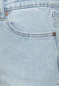 Gaiši zils džinsa audums ar mazu kabatu, kurai ir vara kniedes detalizācija un redzamā šuvuma, radot gludu tekstūru un ikdienišķu izskatu.