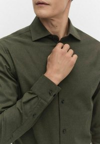 Tumma vihreä nappikauluspaita, jossa on rakenteellinen kaulus, pitkät hihat ja mustat napit hihoissa ja etusarakkeessa; tekstuurinen kangas.