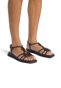 Czarne płaskie sandały z wyszukanymi krzyżującymi się paskami i teksturowaną podeszwą, z regulowanymi klamrami i otwartym noskiem.