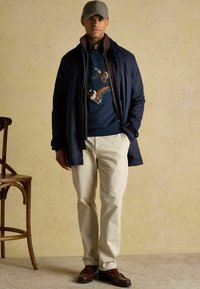 Tumed veekindel jakk pruunide kraega, sinine kampsun kui linnumuster, kreemjad püksid ja pruunid loafersid, neutraalse tausta ees.