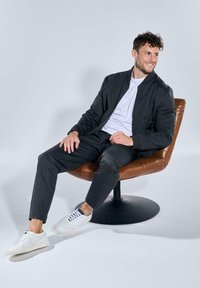 Schwarze Bomberjacke über einem weißen T-Shirt, kombiniert mit schwarzen Hosen und weißen Sneakers. Modell sitzt auf einem braunen Drehstuhl.