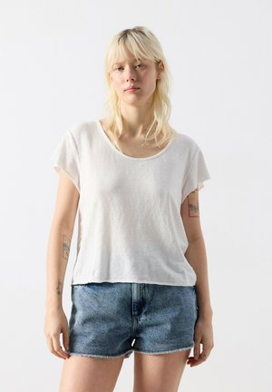 Młoda kobieta o blond włosach ubrana w białą luźną koszulkę i niebieskie denimowe szorty, stoi na tle jednolitego białego tła.