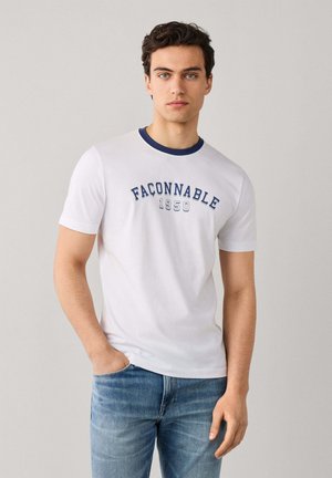 Jeune homme aux cheveux foncés portant un t-shirt blanc Façonnable 1950 et un jean bleu, debout avec une main dans la poche devant un fond uni.