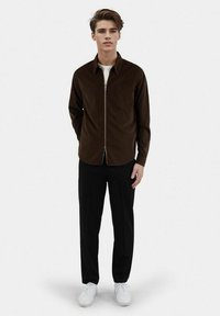 Brown corduroy zip-up shirt with a pointed collar, paired with black trousers and white sneakers, creating a modern casual look.