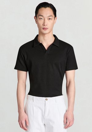 Mann mit schwarzem kurzärmligem Polohemd, das in weiße Hosen mit Nadelstreifen gesteckt ist, steht vor einem schlichten hellen Hintergrund.