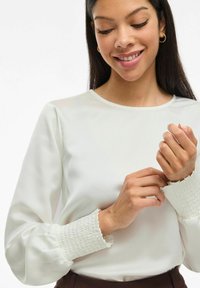 Kvinna som justerar ärmen på sin vita blus med smockade manschetter, ler försiktigt med slutna ögon mot en enkel bakgrund.