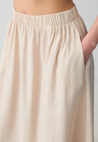 Jupe beige clair avec taille élastique, présentant une texture lisse et des poches latérales, tombant doucement. Pas de motifs ni d'ornements visibles.