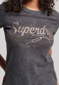 Tummanharmaa t-paita, jossa on kulunut pinta ja jossa lukee "Superdry Merchandise" paksulla fontilla sekä grafiikka mikrofonikaapelista.