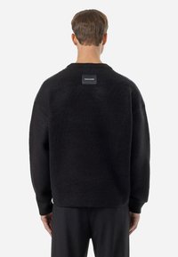 Sweater preto oversized com uma superfície texturizada, apresentando uma etiqueta nas costas com o logótipo "PEGADOR" e detalhes laterais contrastantes.