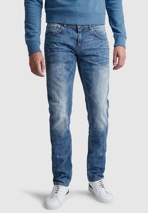 Homme portant un jean bleu délavé, des baskets blanches et un sweat-shirt bleu, debout devant un fond uni.