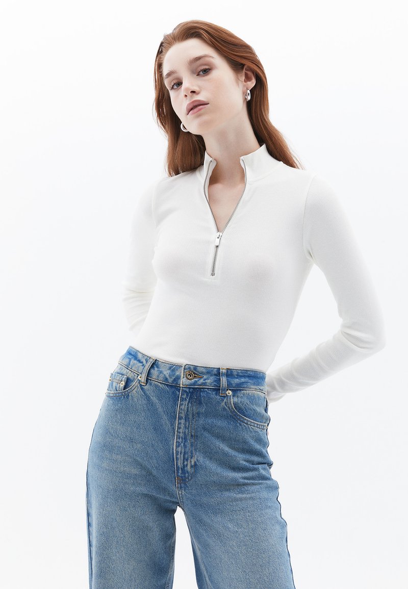Haut à manches longues zippé blanc en texture côtelée, associé à un jean en denim bleu clair taille haute avec un délavage léger et des poches standard.
