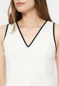 Femme portant un haut blanc sans manches avec une bordure noire le long du col en V et des emmanchures, visible du cou jusqu'au milieu du torse.