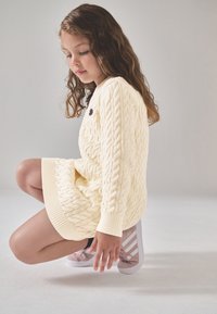 Šviesiai geltona megzta megztinio suknelė su ribsju rankogaliais, derinama su rožiniais sportbačiais, puoštas baltais dryžiais. Minkšta tekstūra, elegantiškas dizainas.