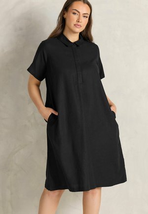 Femme portant une robe courte noire ample à manches courtes avec col et boutons cachés, debout les mains dans les poches sur un fond uni.