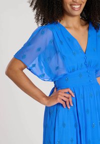 Blå klänning med korta, genomskinliga ärmar, prydd med dekorativ pärlbroderi i diamantformer. Knappar i detalj längs framsidan, flödande tygtextur.