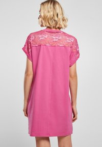Roze jurk met kanten details op de rug en korte mouwen, met een losse pasvorm die tot halverwege de dij reikt. Zachte textuur door heel de jurk.