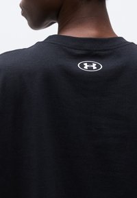 Κοντινή λήψη του άνω μέρους της πλάτης ενός ατόμου που φορά ένα μαύρο μπλουζάκι Under Armour με το λευκό λογότυπο στο κέντρο κάτω από το λαιμό.