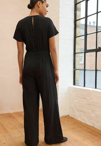 Schwarzer Jumpsuit mit kurzen Ärmeln, lockerer Passform, elastischem Bund, weitem Bein design und einem Knopfverschluss hinten. Strukturiertes Stoffdetail.