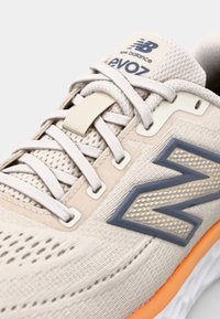 Beige sportsko med en tekstureret overdel, lysegrå snørebånd, fremtrædende blåt logo og en orange detalje på sålen.