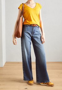 Geel V-hals T-shirt, blauwe wijde denim jeans, bruine riem, bruine totebag en oranje-gele schoenen met witte strepen. Platte houten vloer als achtergrond.