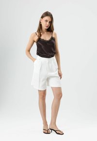 Zwarte satijnen camisole met kanten afwerking, gecombineerd met witte geplooide shorts. Het model draagt zwarte teenslippers, gestyled tegen een neutrale achtergrond.
