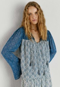 Vrouw met golvend blond haar draagt een blouse met lange mouwen met een blauw patroon, crème en blauw geometrisch dessin en kwastjes, staand tegen een muur.