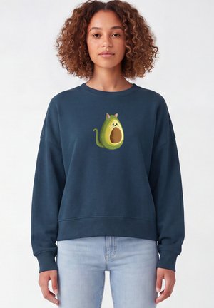 Jonge vrouw met krullend haar die een marineblauwe trui draagt met een groene avocado kat-afbeelding aan de voorkant.